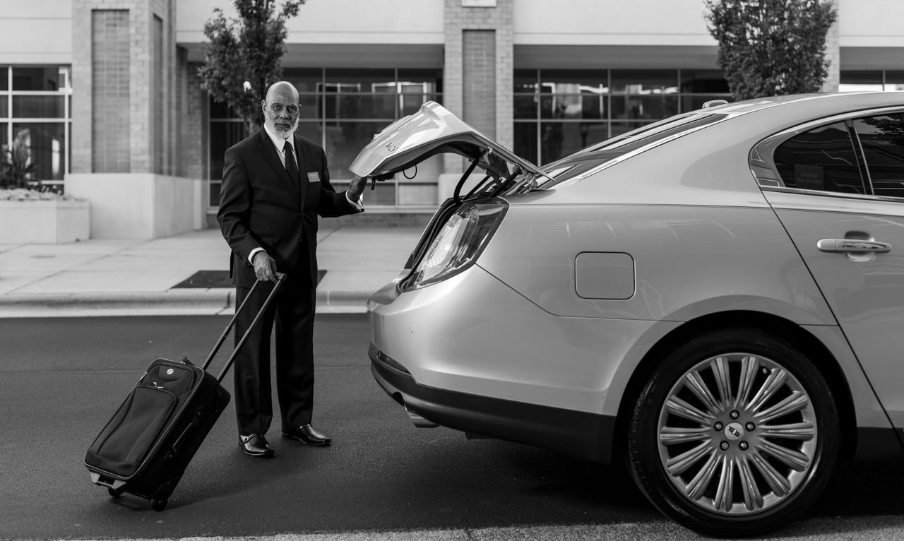Chauffeur in suit loading luggage into luxury car trunk outside a modern building.