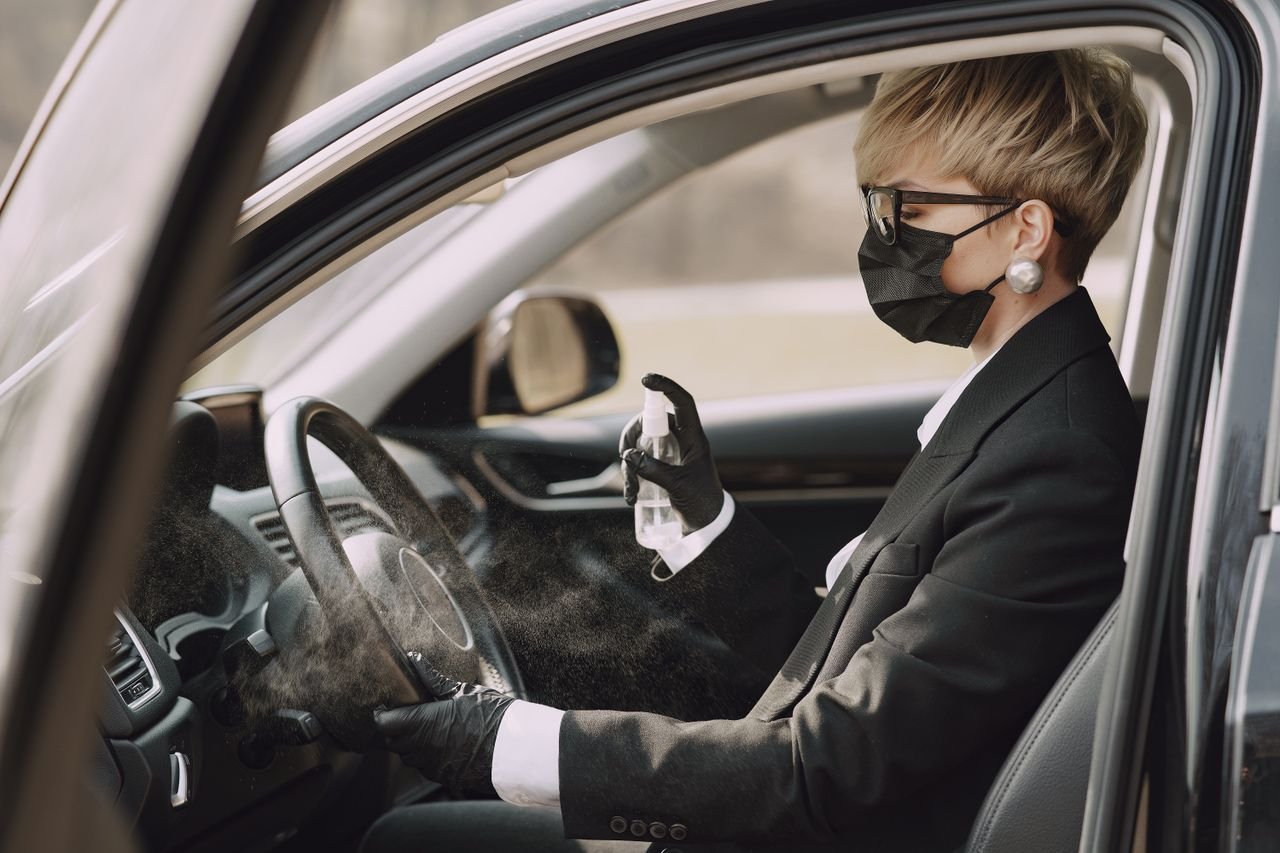 Side view of businesswoman in elegant suit and protective mask with gloves sitting on driver seat and spraying sanitizer on wheel