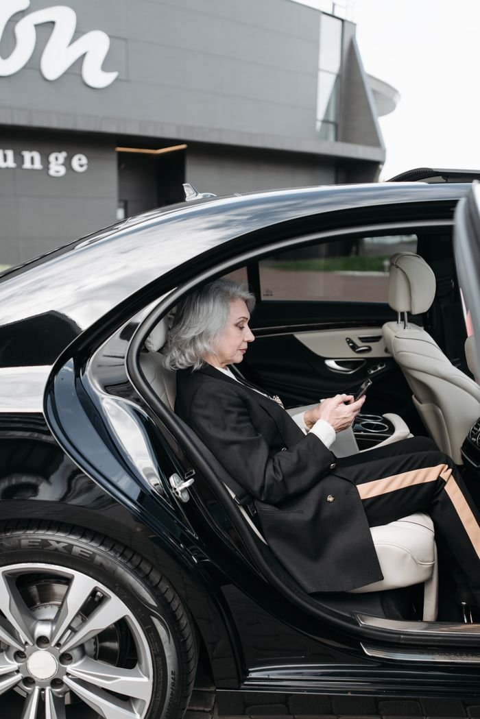 Senior woman in business attire sits in a luxury car, using a tablet outside a modern building.