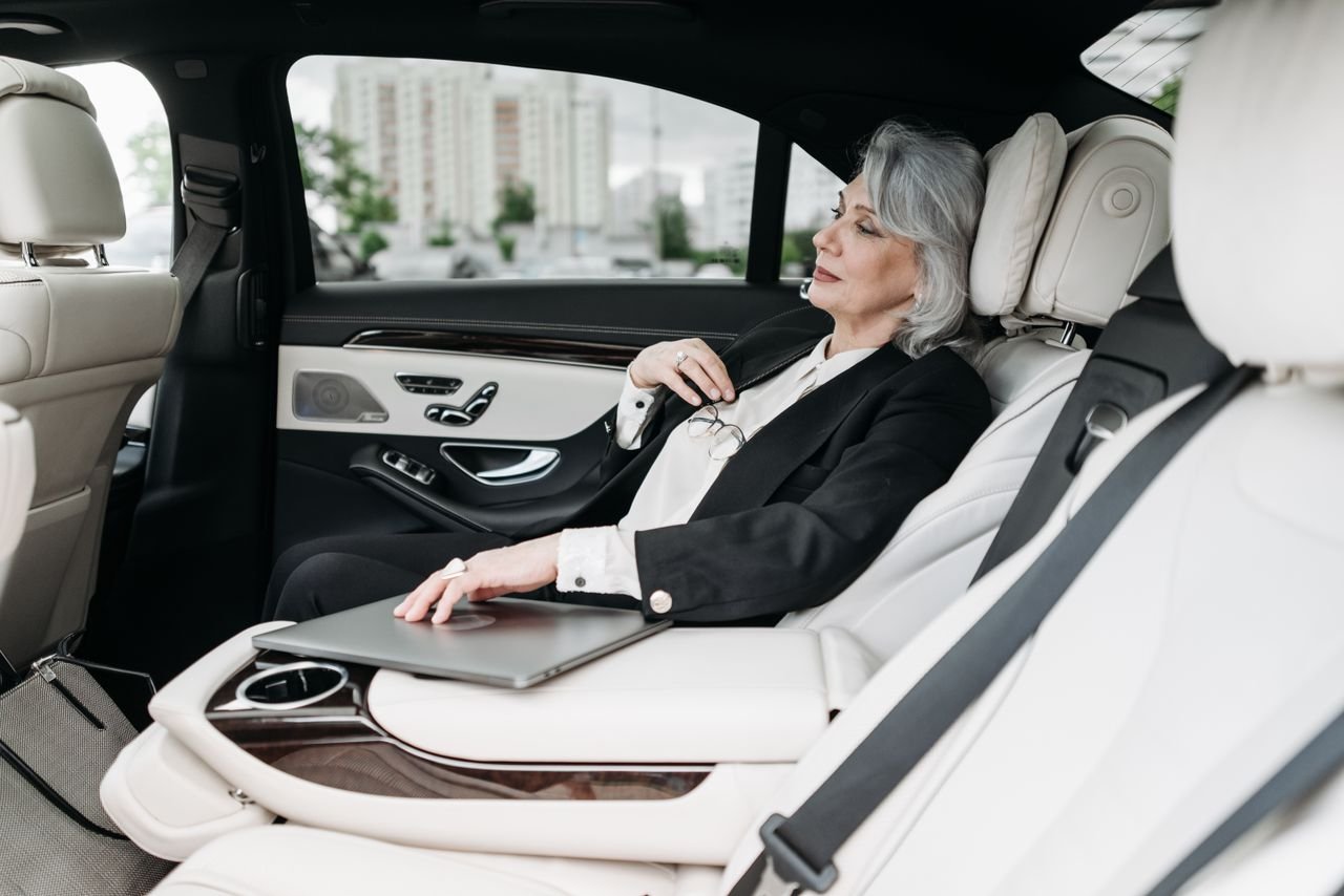 Senior woman in a luxury car with a laptop, gazing out the window, representing business travel.