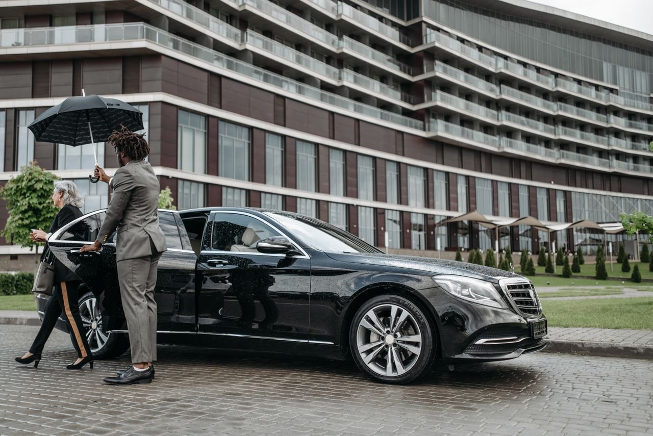 A business professional exits a luxury car while being shielded by an umbrella.