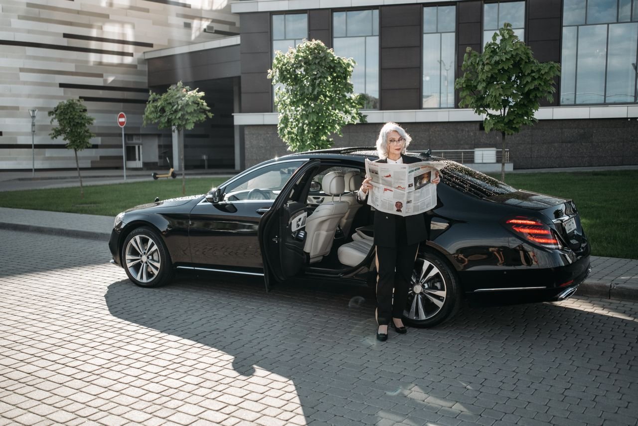 Senior woman reading a newspaper beside a luxury car parked outside a modern building.